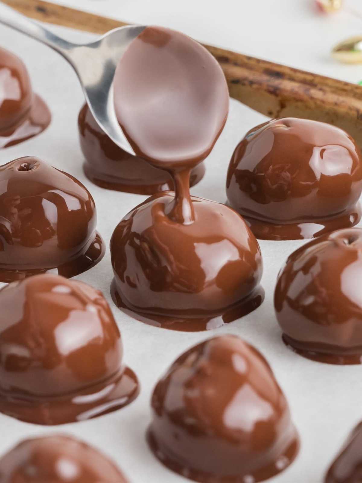 A spoon pours melted chocolate over round candies on a parchment-lined baking sheet.