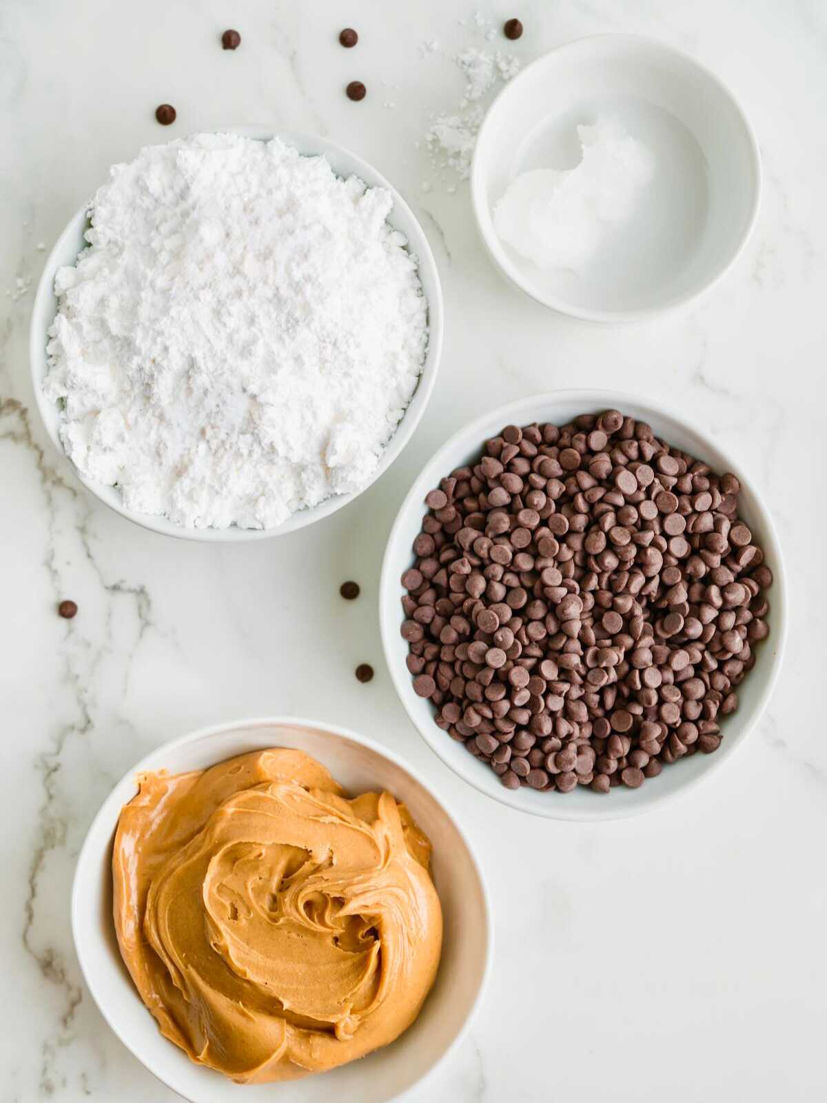 Four bowls with powdered sugar, mini chocolate chips, coconut oil, and creamy peanut butter on a white surface.