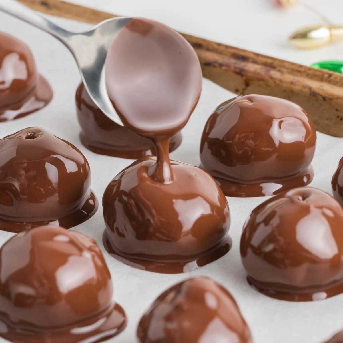 A spoon drizzles melted chocolate over round chocolate-covered peanut butter balls on parchment paper.