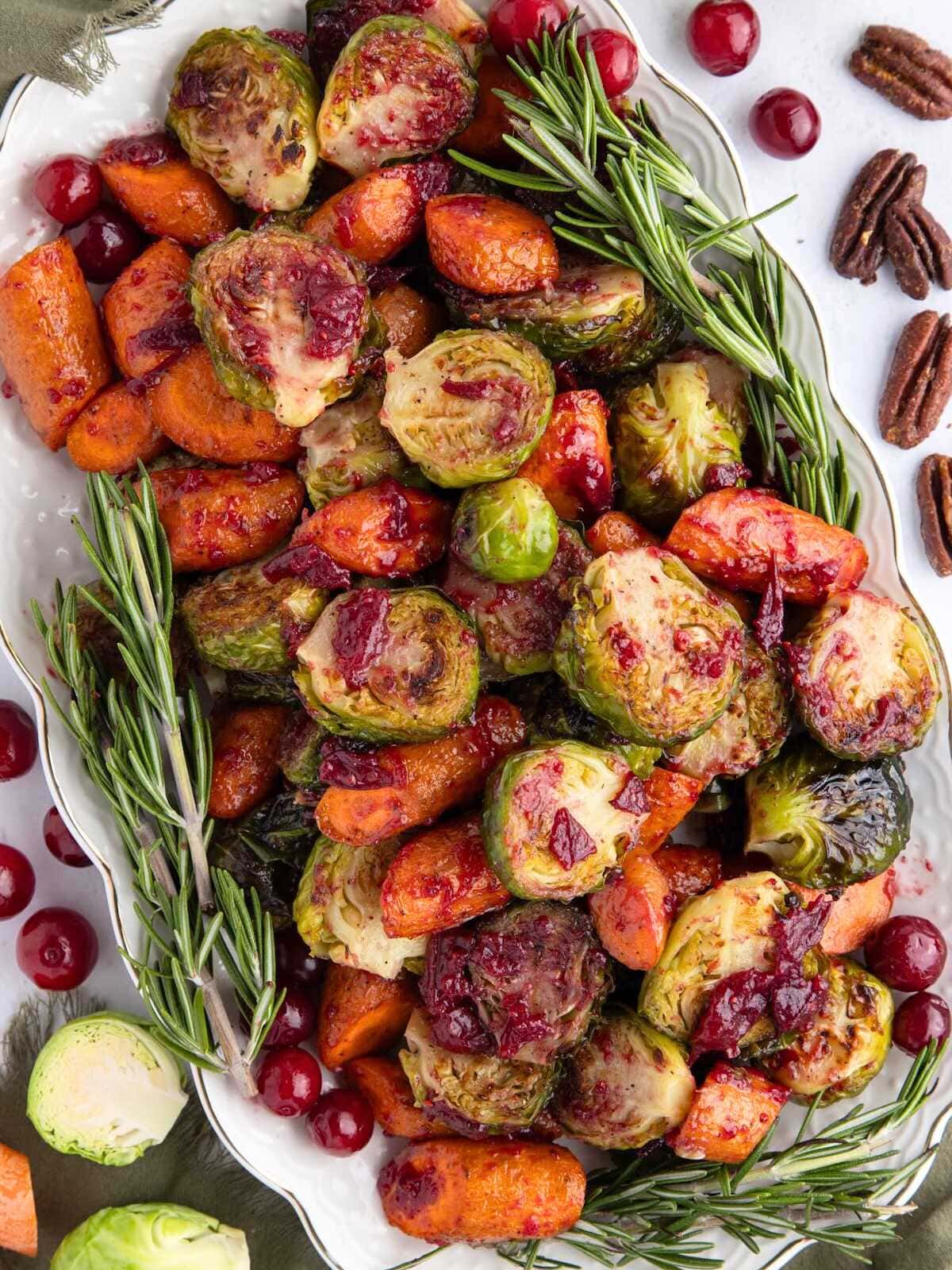 Roasted Brussels sprouts and carrots with cranberries and rosemary on a white platter, garnished with pecans.