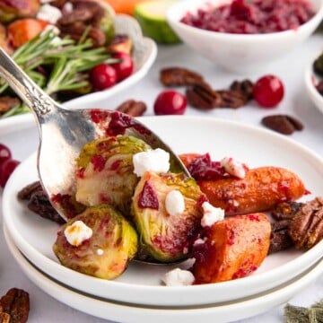 A spoon serves Brussels sprouts, carrots, goat cheese, and cranberry sauce onto a white plate with pecans.