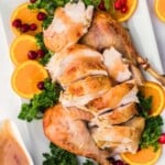 Sliced roasted turkey with gravy, garnished with orange slices, parsley, and cranberries on a white platter.