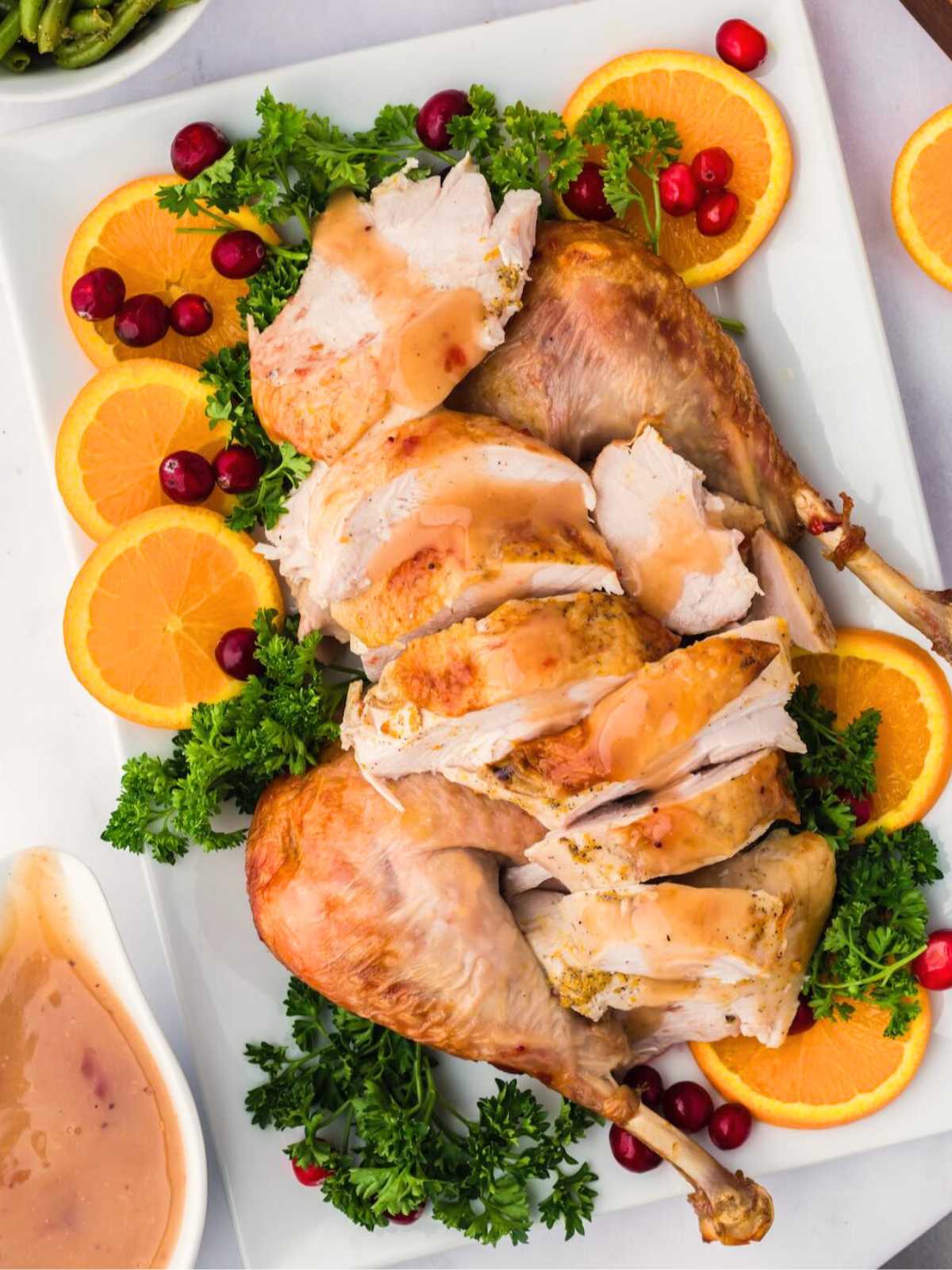 Sliced roasted turkey with gravy, garnished with orange slices, parsley, and cranberries on a white platter.
