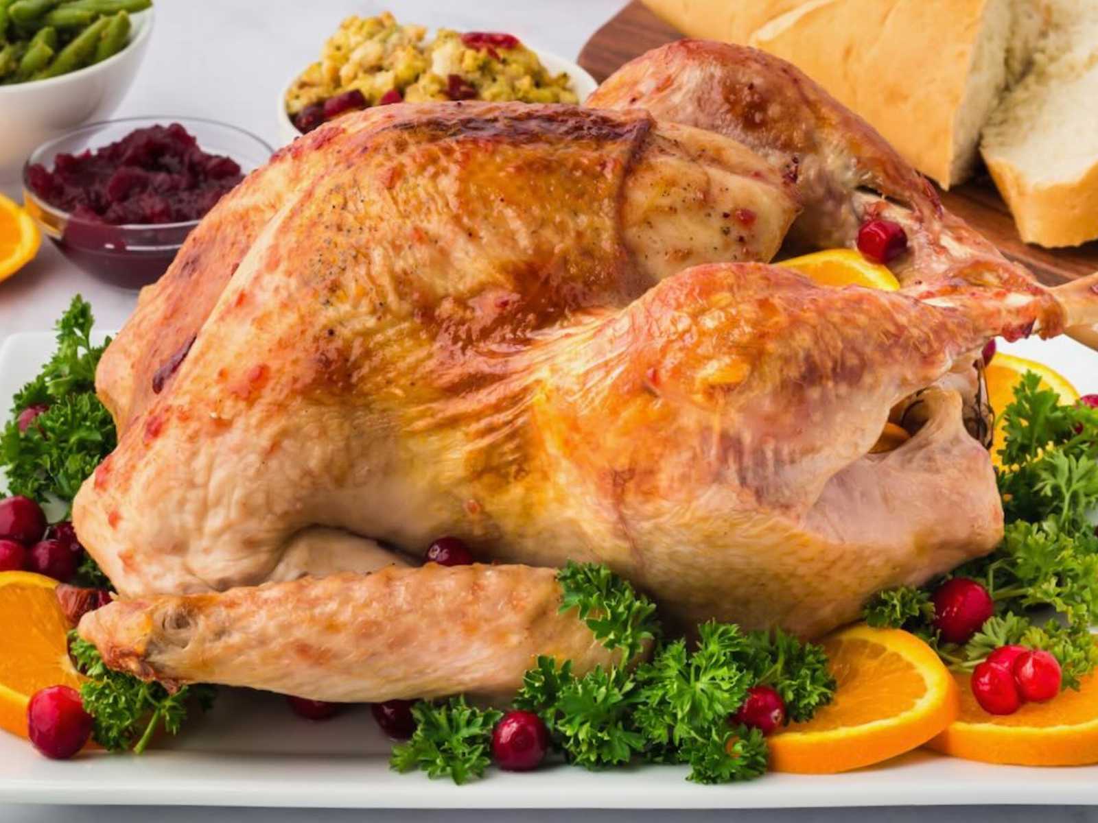 Roast turkey on a platter with orange slices, cranberries, and parsley, surrounded by bread and side dishes.