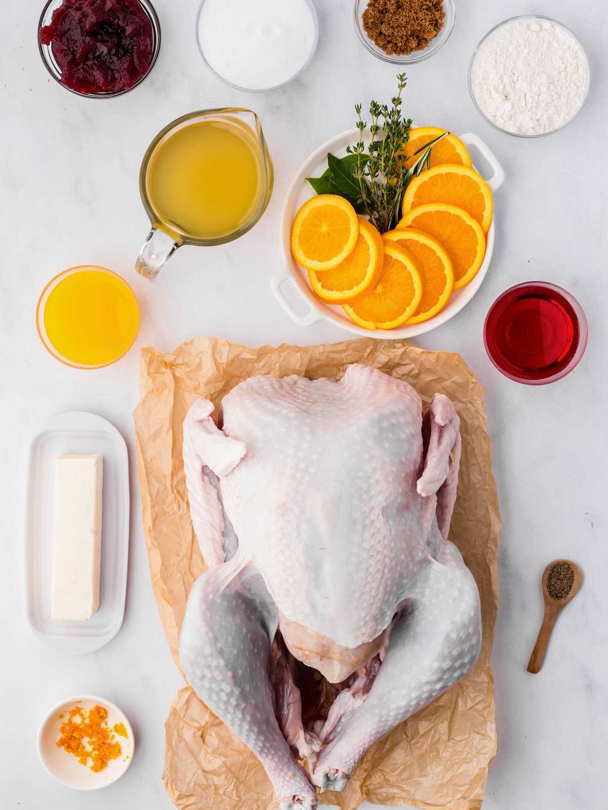 Raw whole turkey on parchment, surrounded by orange slices, herbs, butter, juices, flour, sugar, and seasonings.