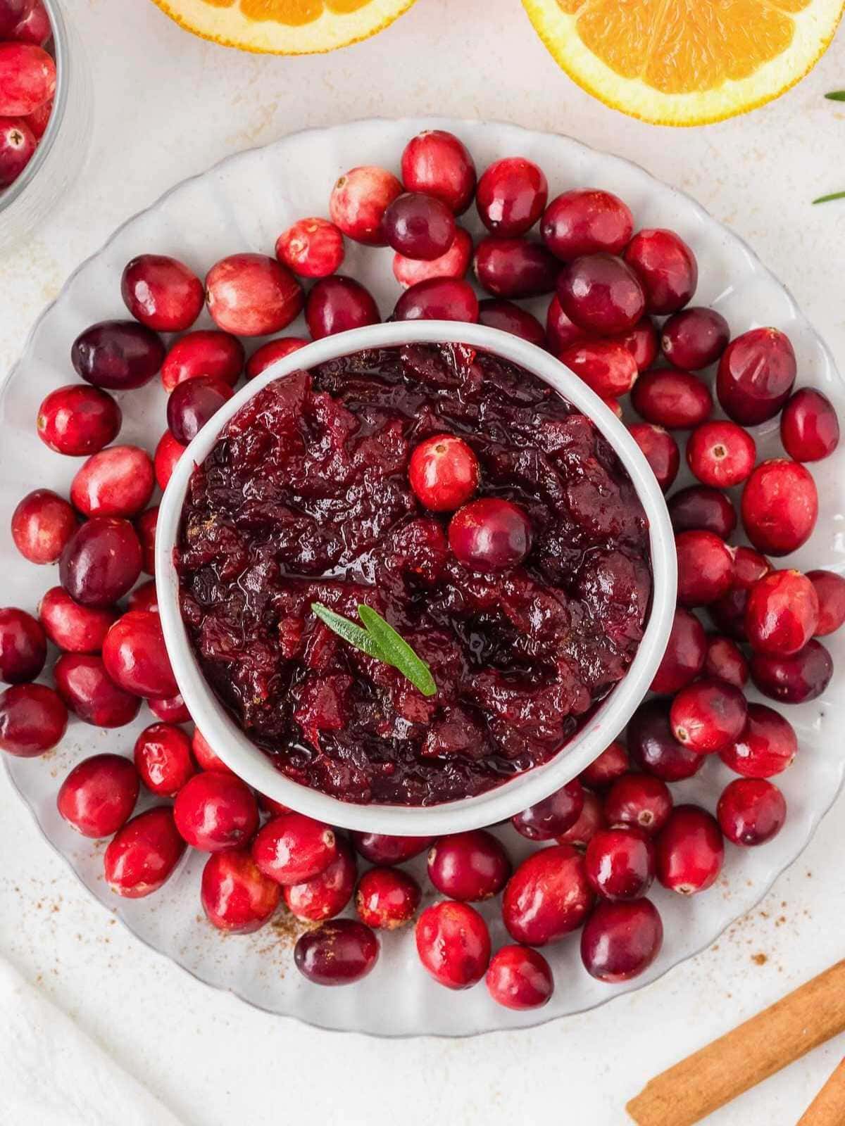 A bowl of cranberry sauce is surrounded by fresh cranberries on a white plate, with orange slices nearby.