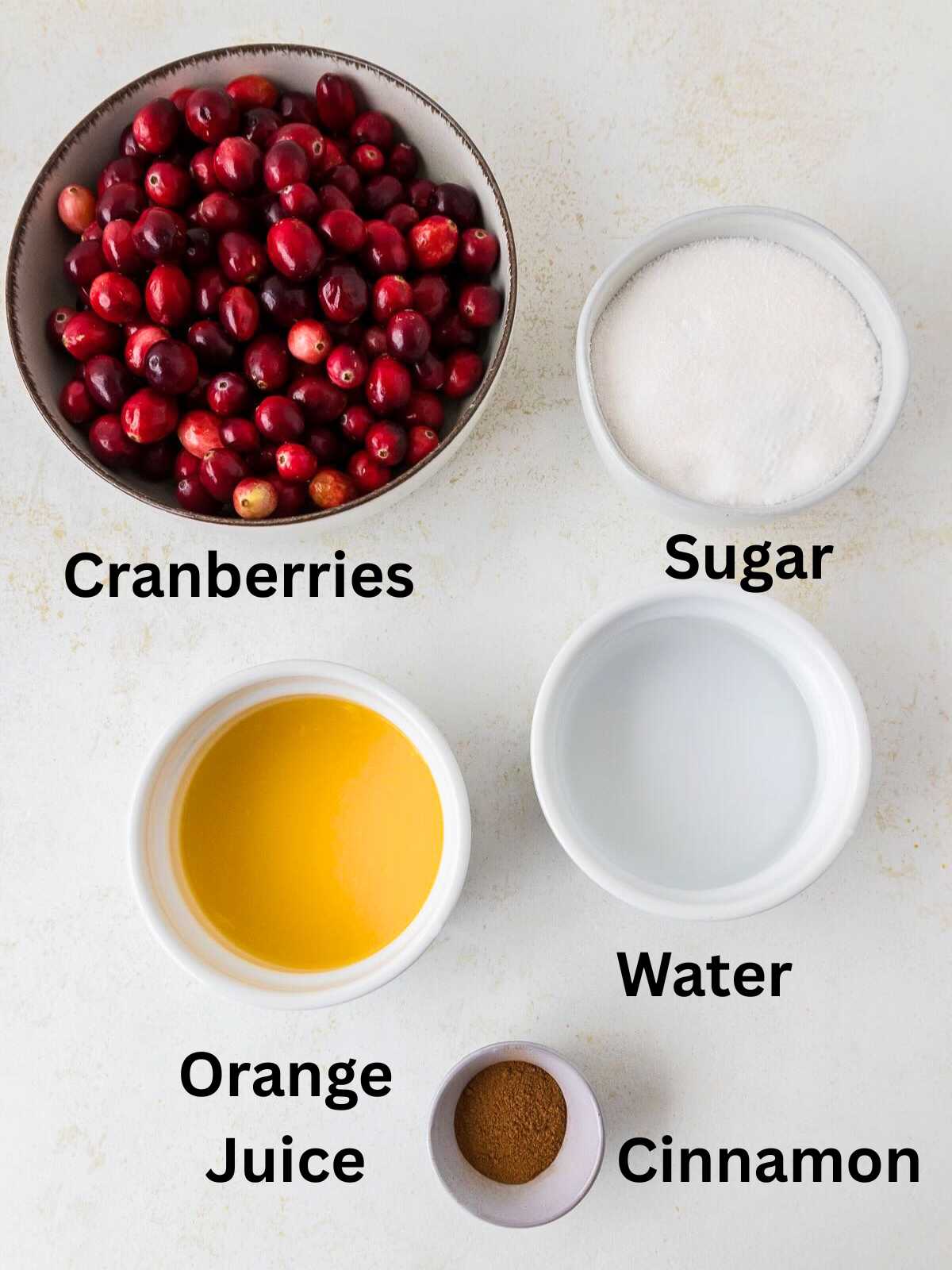 Five labeled bowls containing cranberries, sugar, orange juice, water, and cinnamon on a white surface.