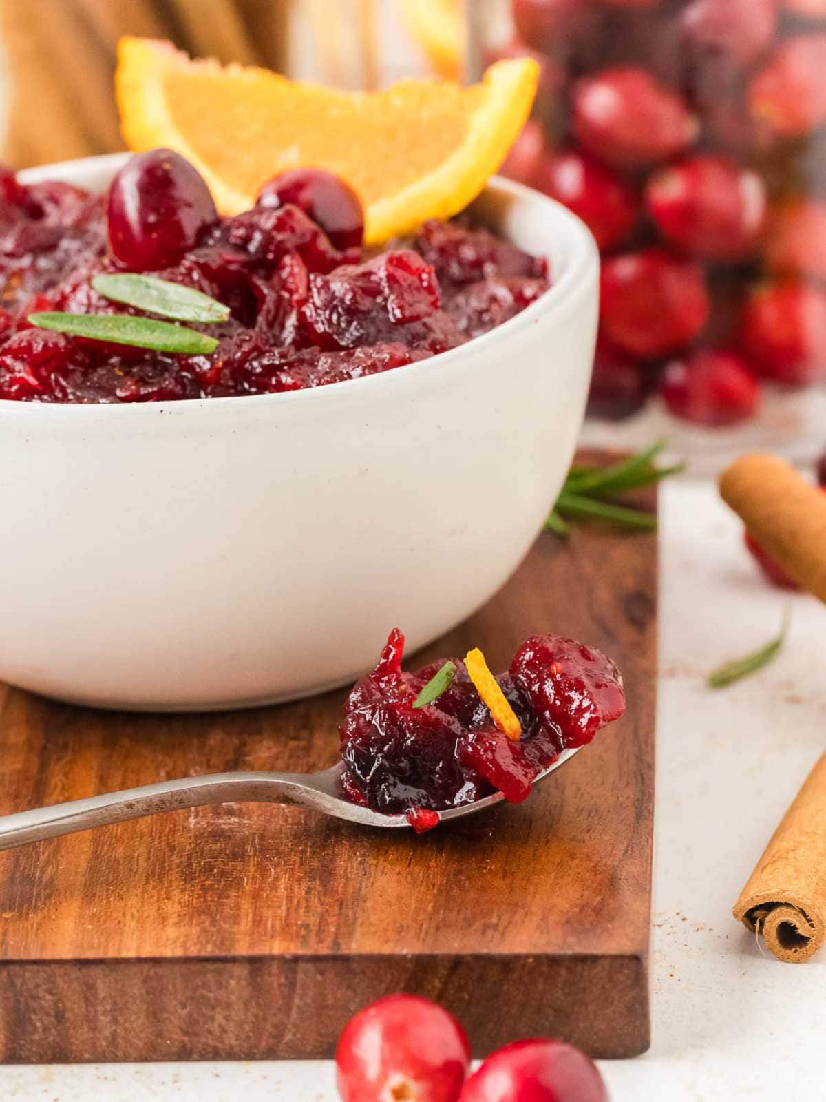 Bowl of cranberry sauce with orange slice and rosemary, with a spoonful in front on a wooden board.