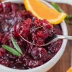 Spoonful of whole berry cranberry sauce above a bowl, garnished with orange slice and green herbs.