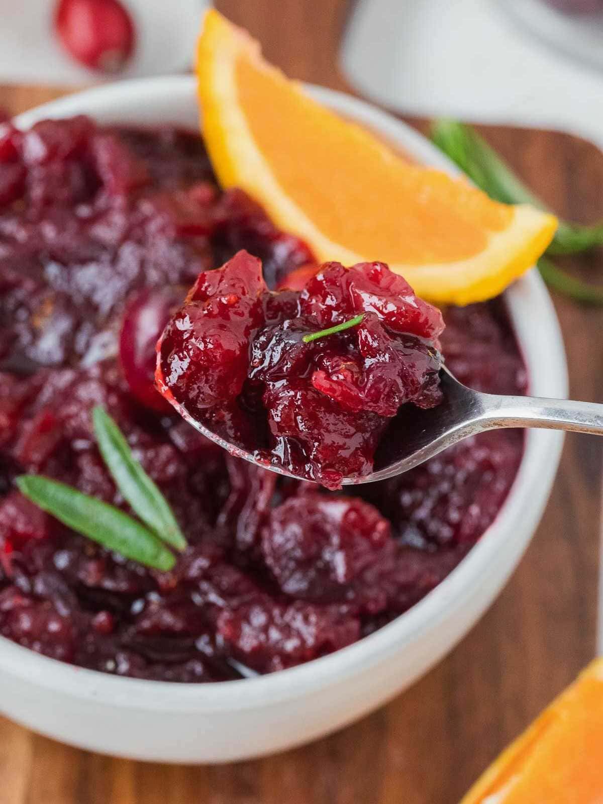 Spoonful of whole berry cranberry sauce above a bowl, garnished with orange slice and green herbs.