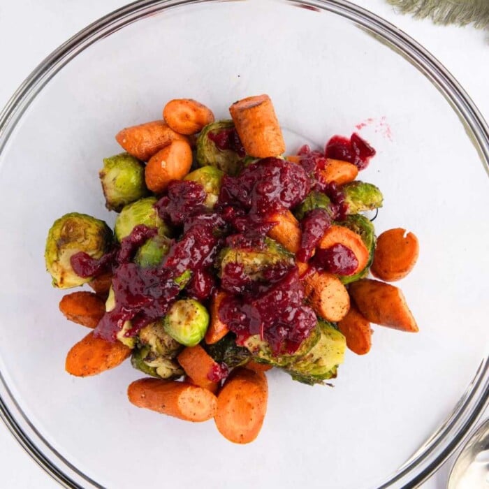Roasted Brussels sprouts and carrots topped with a red cranberry sauce in a clear glass bowl.