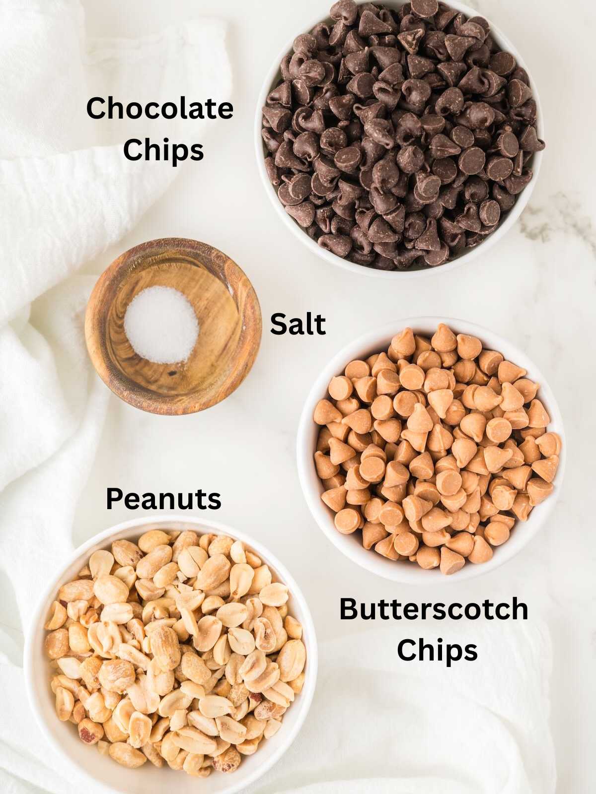 Bowls of chocolate chips, butterscotch chips, peanuts, and a small bowl of salt on a white surface.