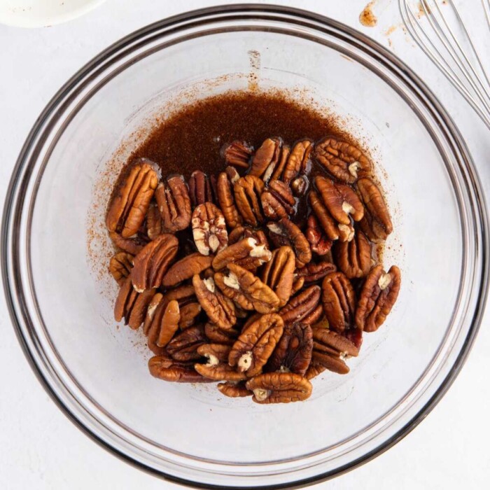 Pecans with spices in a glass mixing bowl on a white surface.