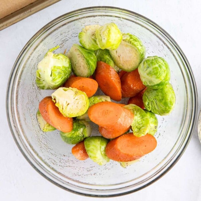 Sliced carrots and halved Brussels sprouts in a clear glass bowl, seasoned and ready to cook.