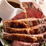 Sliced prime rib on a plate with gravy being poured over it, garnished with fresh rosemary.