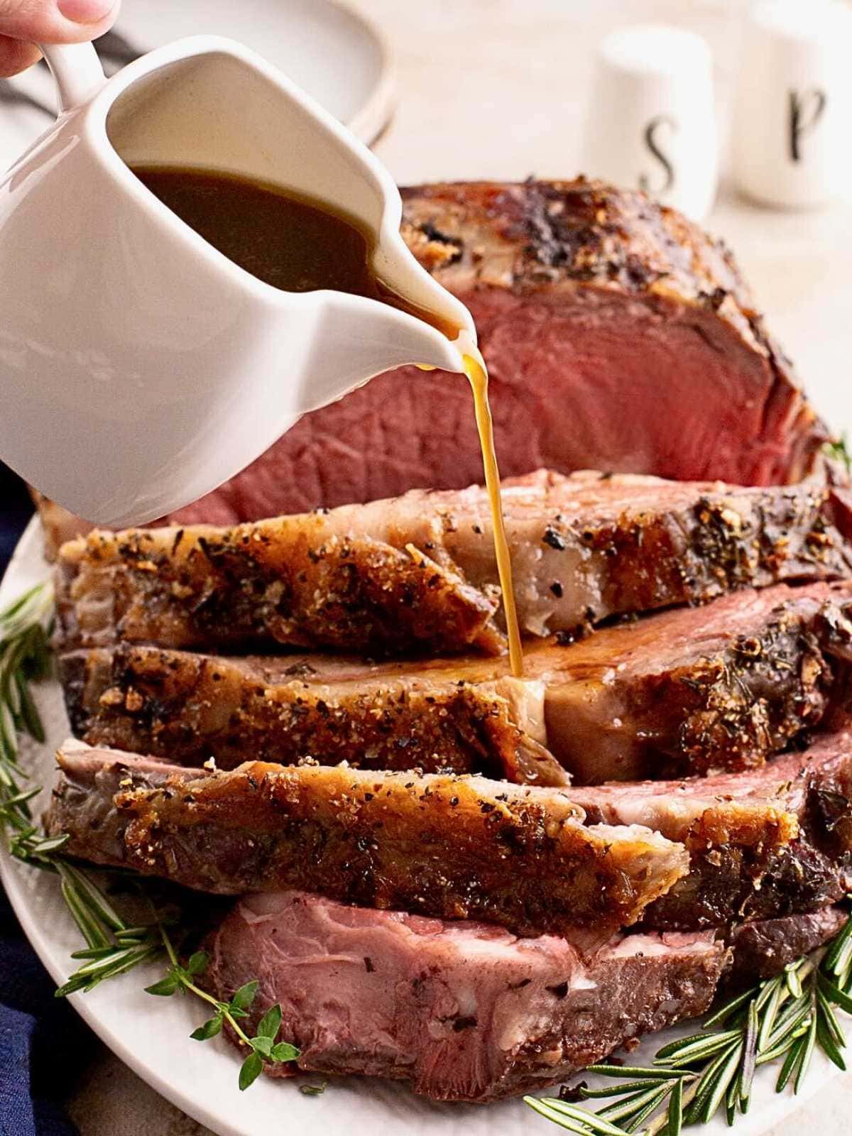 Sliced prime rib on a plate with gravy being poured over it, garnished with fresh rosemary.