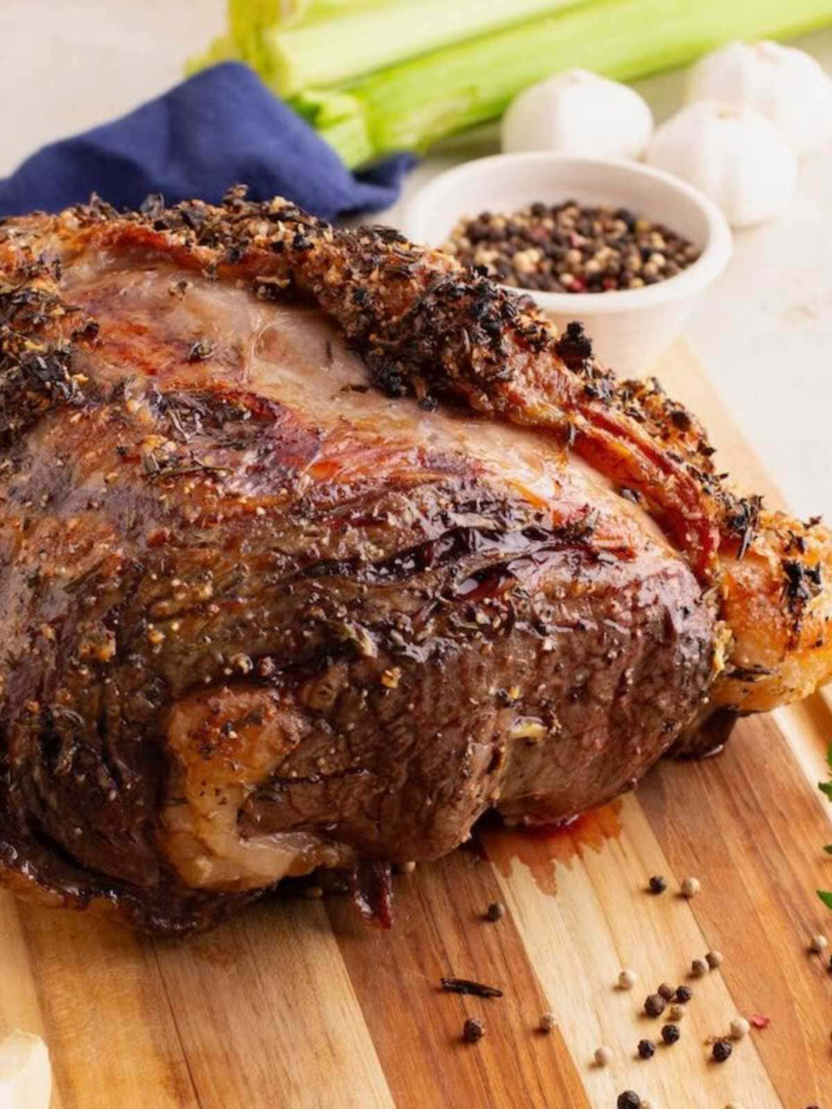 Prime rib with herbs on a wooden cutting board, peppercorns scattered, celery and garlic in the background.