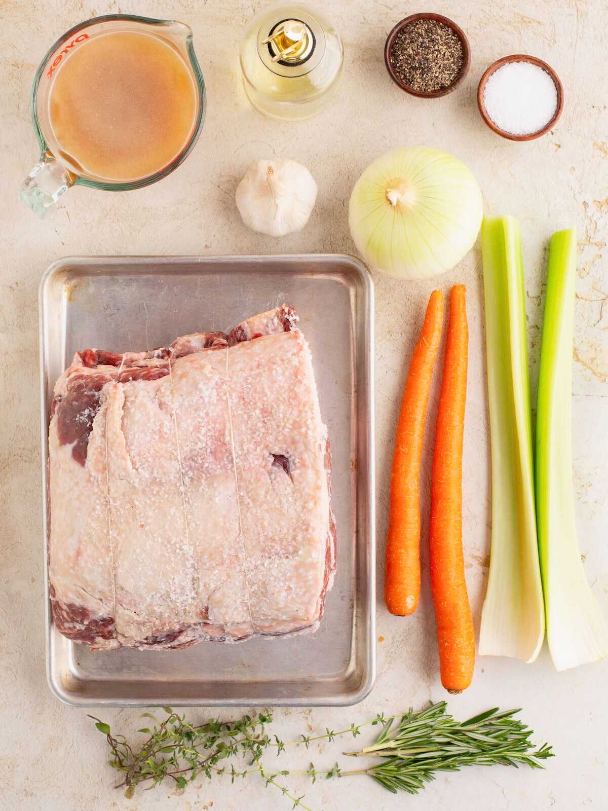 Raw beef roast on a tray, with carrots, celery, onion, garlic, herbs, broth, oil, salt, and pepper beside it.