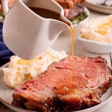 Gravy being poured onto a slice of prime rib with mashed potatoes on a plate in the background.