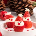 Three strawberry Santa treats with cream and chocolate chip faces on a white plate, surrounded by pinecones.
