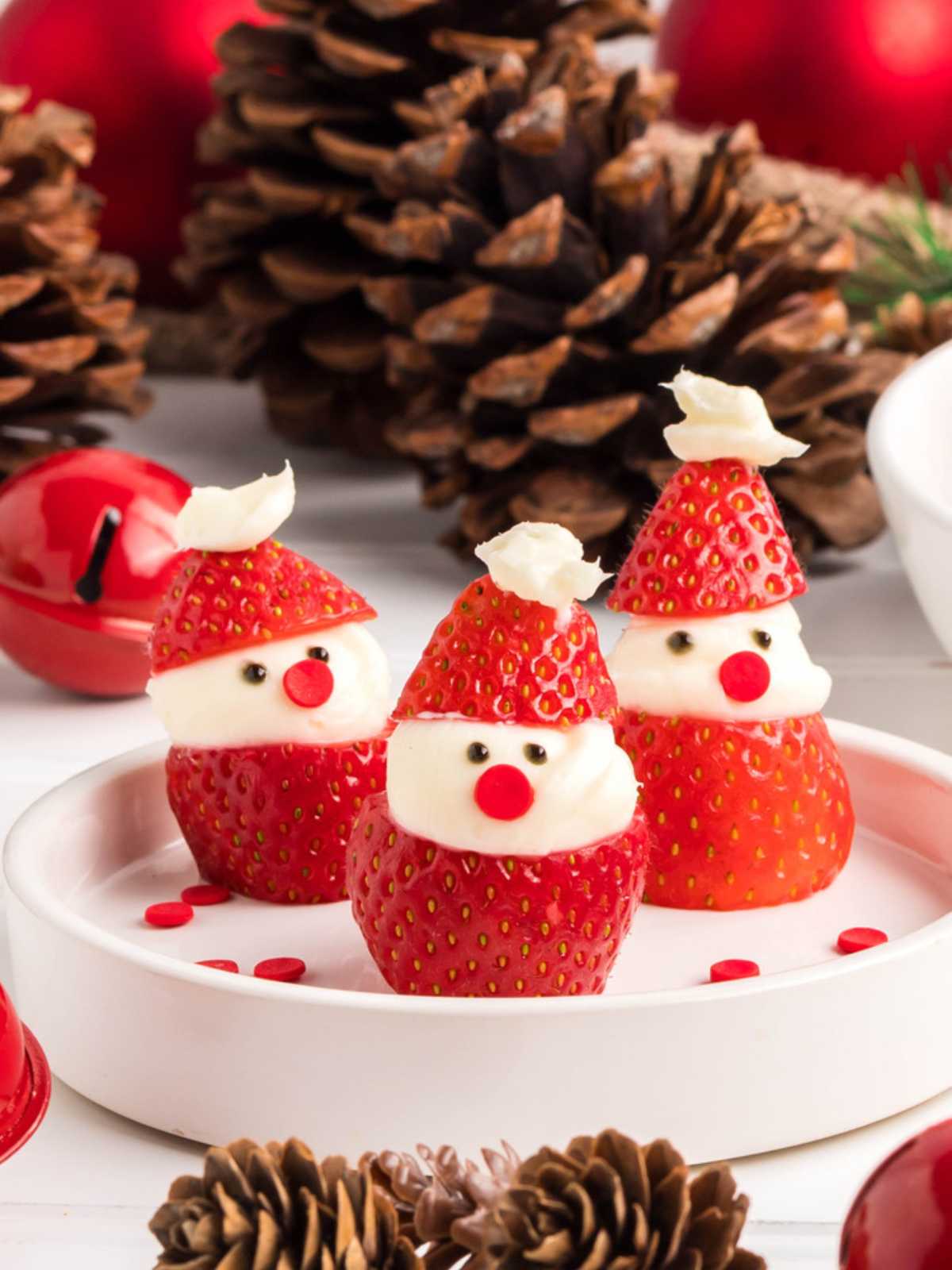 Three strawberry Santa treats with cream and chocolate chip faces on a white plate, surrounded by pinecones.