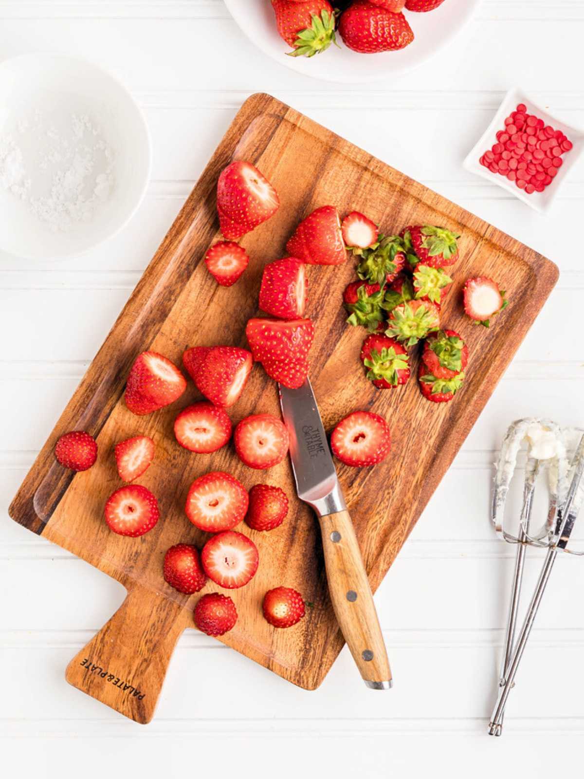 Sliced strawberries and a knife on a wooden cutting board, with bowls of strawberries and ingredients nearby.
