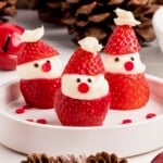 Three strawberries decorated as mini Santas with cream and candy faces on a white plate.