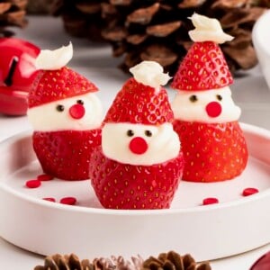Three strawberries decorated as mini Santas with cream and candy faces on a white plate.