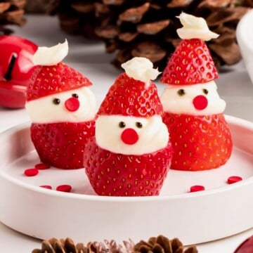 Three strawberries decorated as mini Santas with cream and candy faces on a white plate.