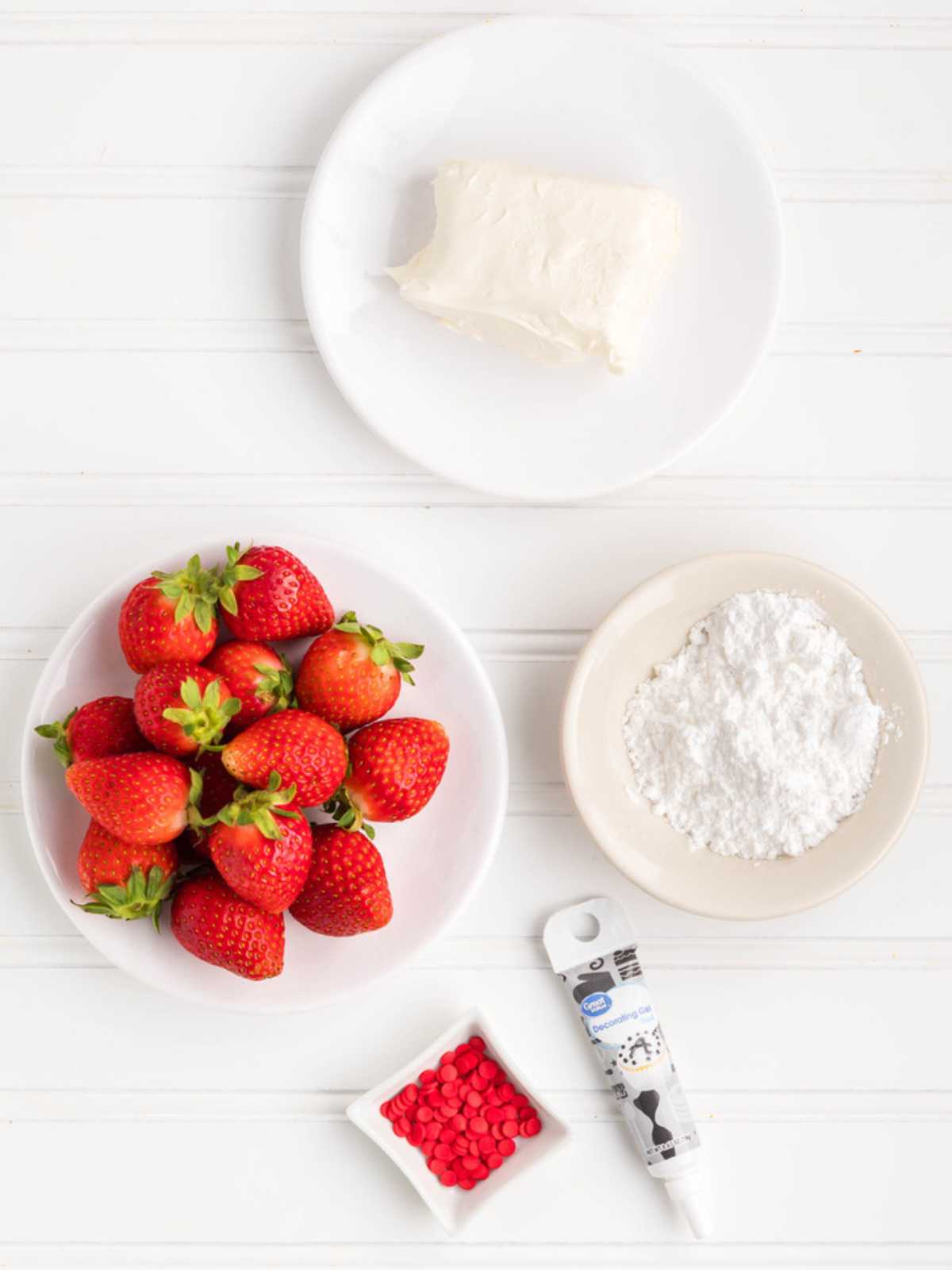 Four bowls with cream cheese, strawberries, powdered sugar, red candy, and a tube of black gel on a white table.
