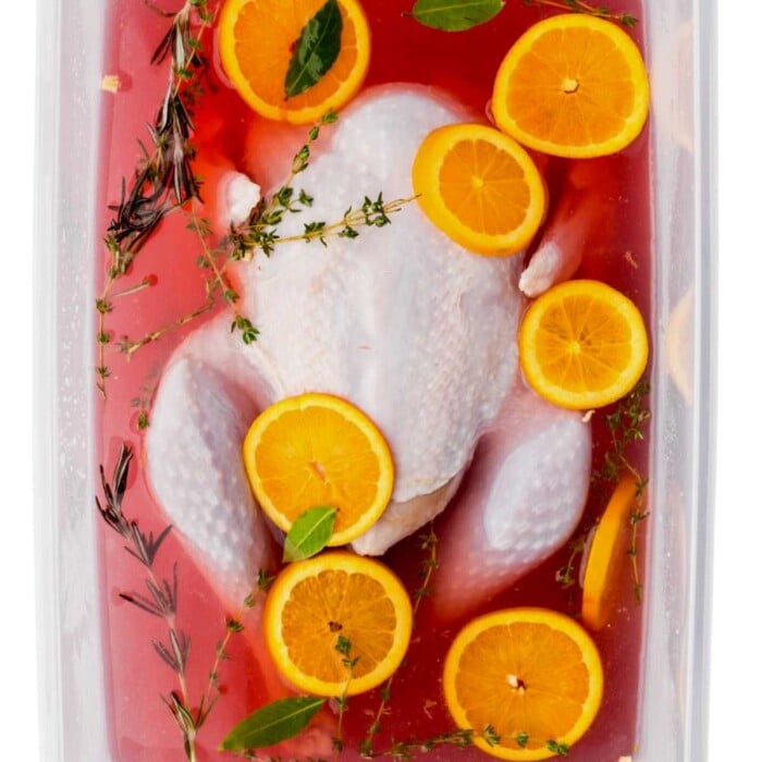 Whole raw chicken in red marinade, topped with orange slices, herbs, and bay leaves in a container.