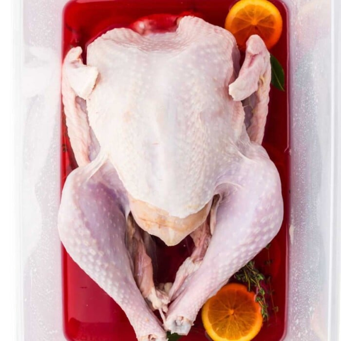 Raw whole turkey in red brine with orange slices and herbs in a clear container, ready for marinating.