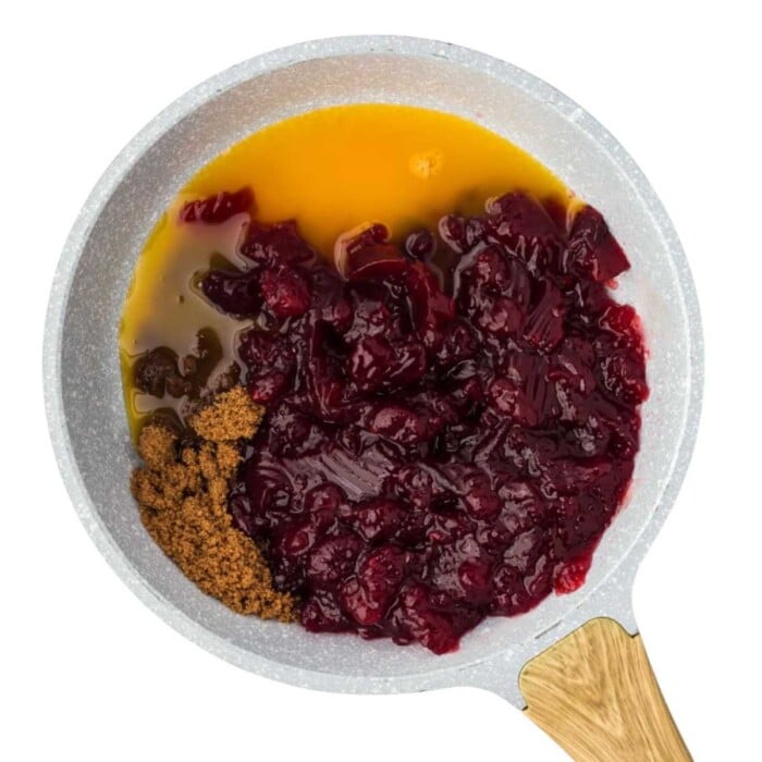 A saucepan with cranberries, orange juice, and brown sugar on a white background.