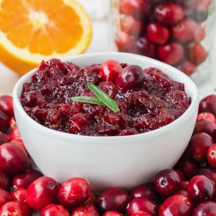 A bowl of cranberry sauce garnished with rosemary, surrounded by fresh cranberries and a sliced orange.