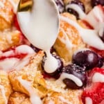 A spoon drizzles white icing over baked bread, blueberries, and strawberries.