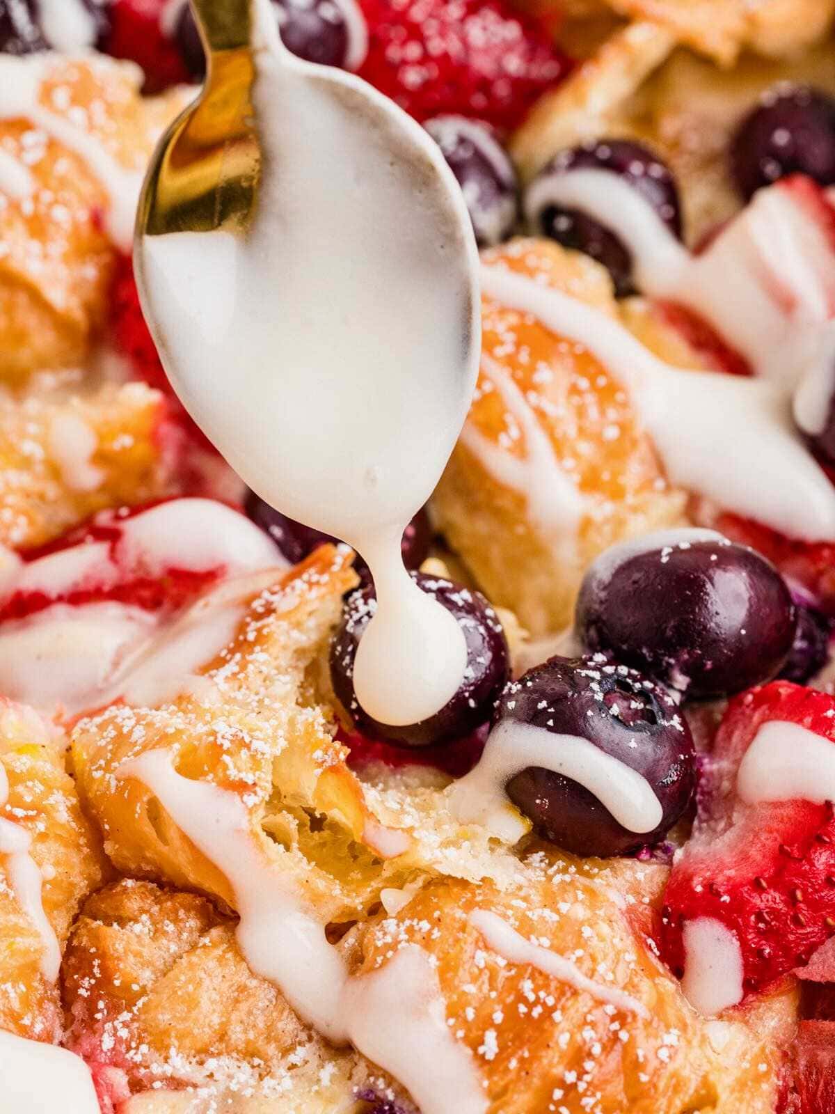 A spoon drizzles white icing over baked bread, blueberries, and strawberries.