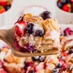 A wooden spatula holds a slice of berry-studded croissant casserole drizzled with icing.