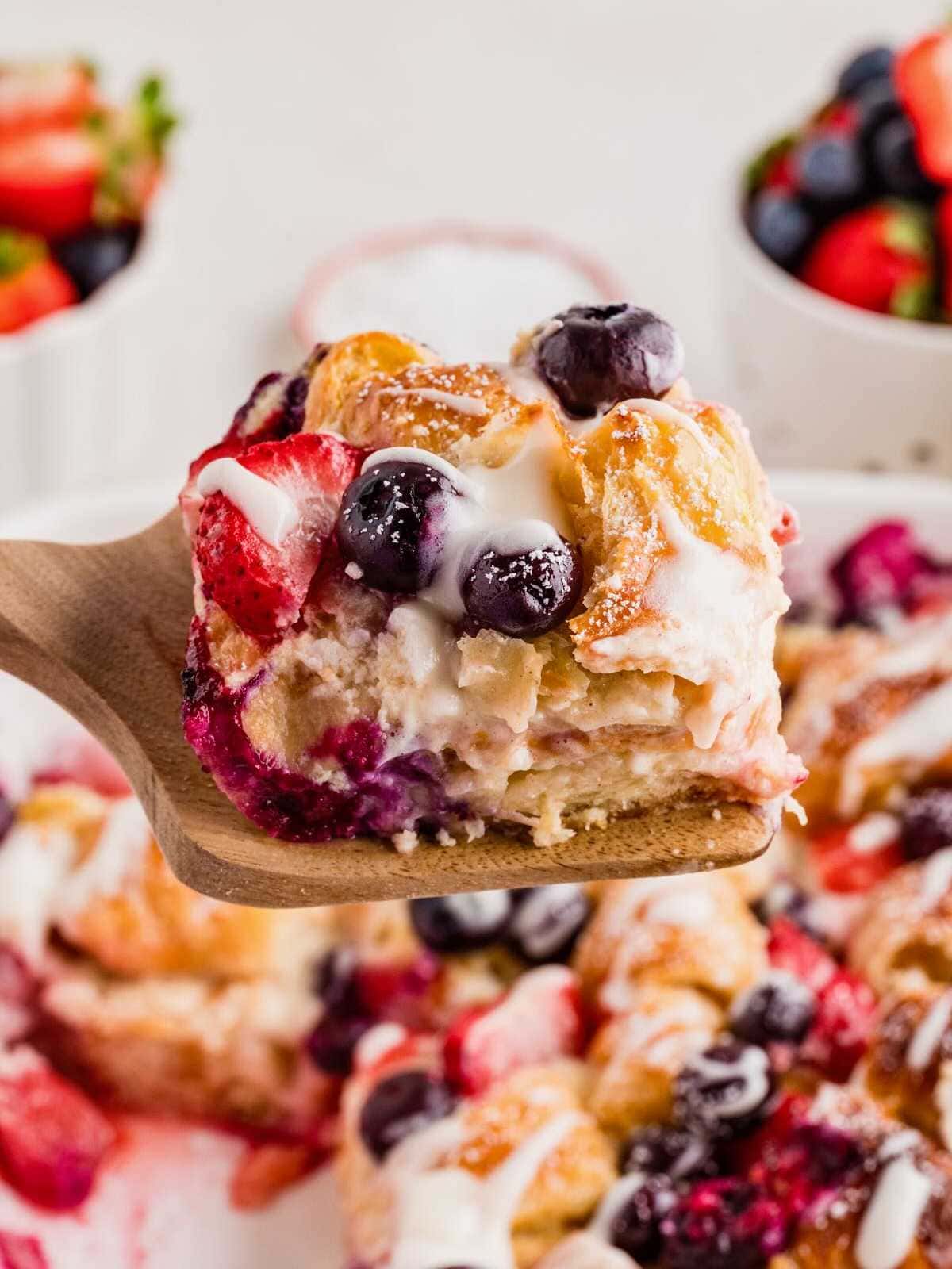 A wooden spatula holds a slice of berry-studded croissant casserole drizzled with icing.