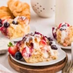 A plate of berry-topped sweet pastry drizzled with icing, with croissants and fresh fruit in the background.