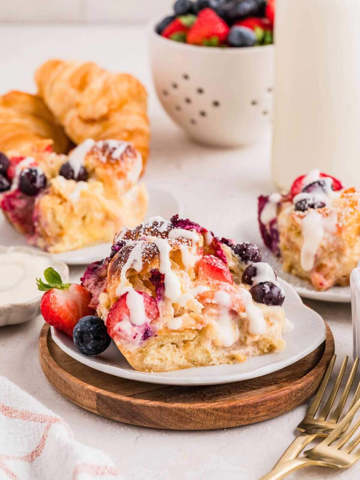 A plate of berry-topped sweet pastry drizzled with icing, with croissants and fresh fruit in the background.
