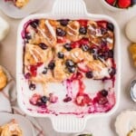 A square dish of berry croissant bake topped with icing, surrounded by fresh berries and served portions.