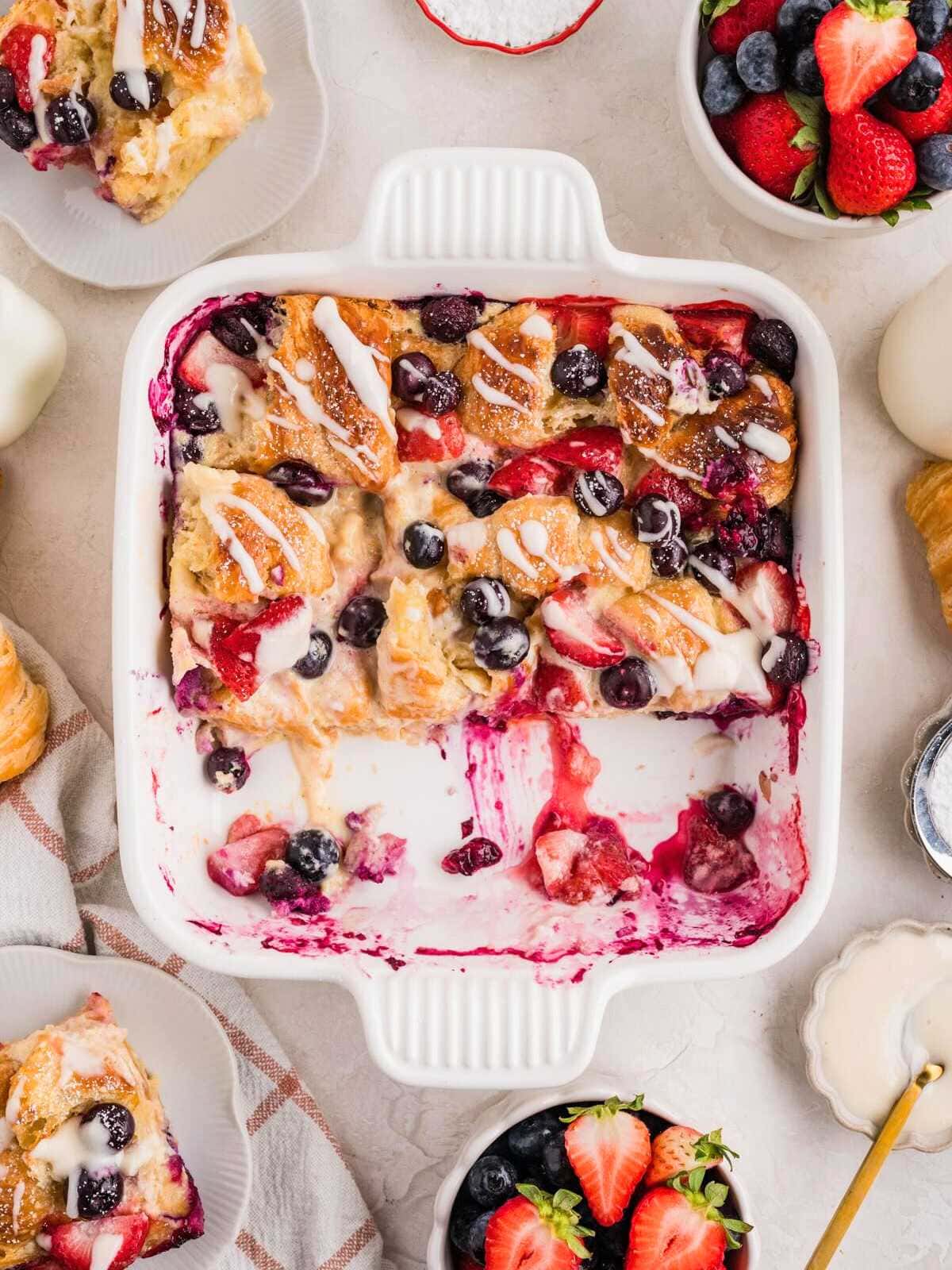 A square dish of berry croissant bake topped with icing, surrounded by fresh berries and served portions.
