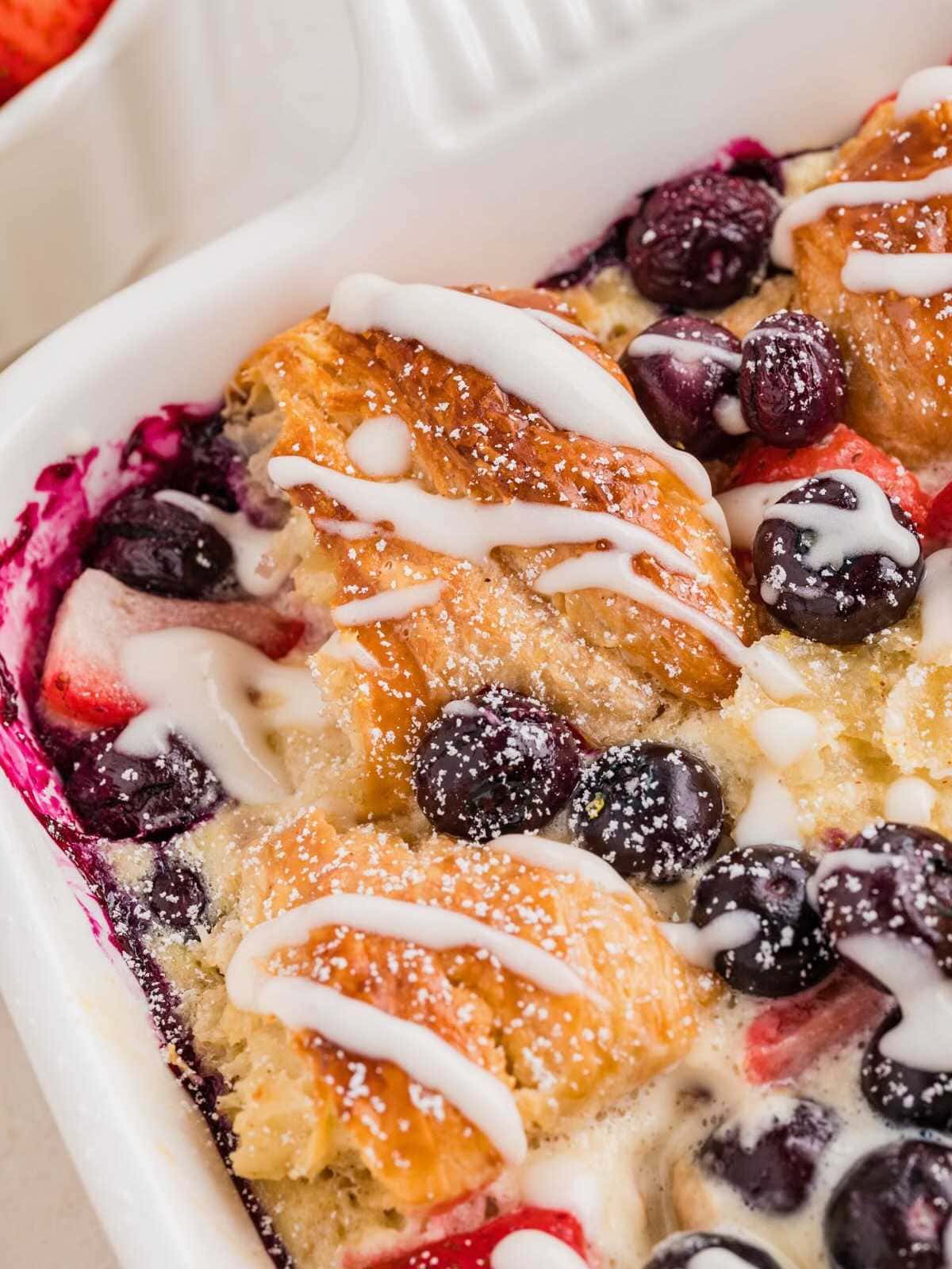 Close-up of a baked pastry casserole with mixed berries and white icing drizzled on top.