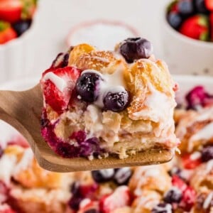 A wooden spatula holds a piece of berry-studded croissant breakfast casserole drizzled with icing.