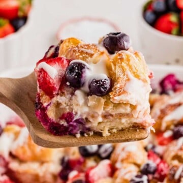 A wooden spatula holds a piece of berry-studded croissant breakfast casserole drizzled with icing.