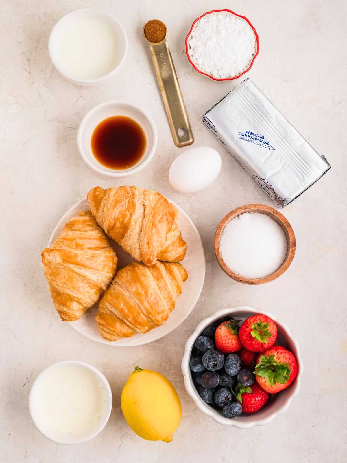 Assorted baking ingredients, croissants, berries, lemon, egg, and dairy on a white surface.