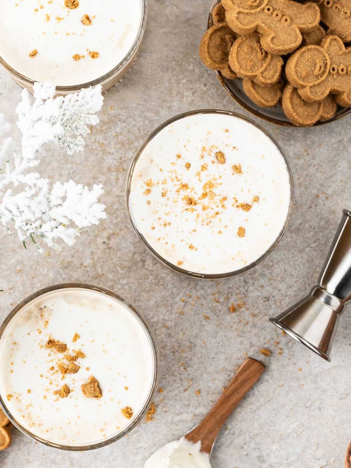 Three creamy drinks topped with cookie crumbs, a plate of cookies, and a jigger on a light countertop.