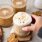 Hand holding a glass of frothy coffee with cookie crumbs, next to two drinks and gingerbread cookies.
