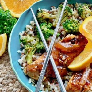 Bowl of rice with broccoli and jerk chicken, garnished with orange slices and metal chopsticks on top.