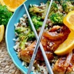 A bowl of rice with broccoli, glazed chicken, orange slices, and metal chopsticks, on a woven placemat.
